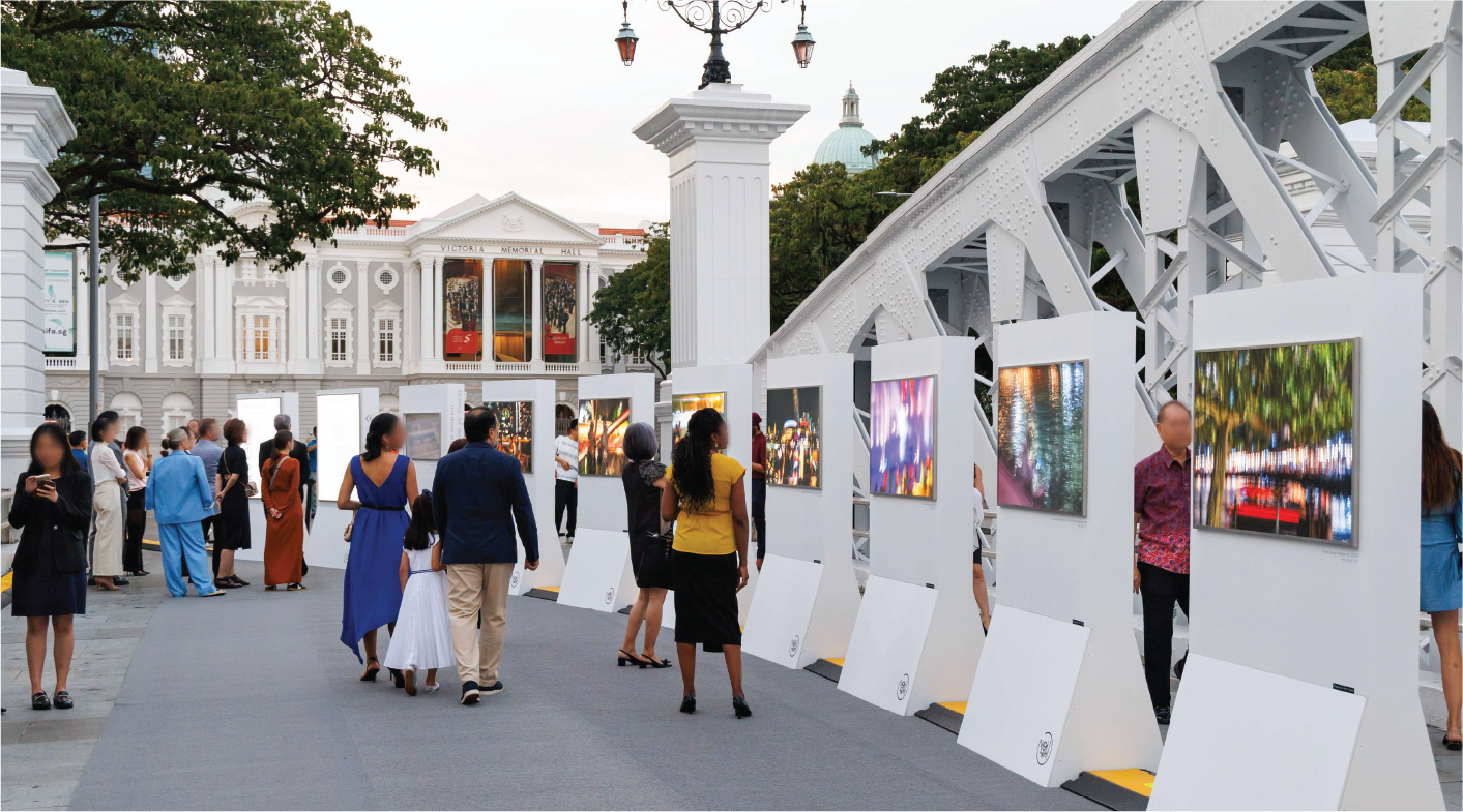 People viewing an outdoor photography exhibit near a white bridge and building.